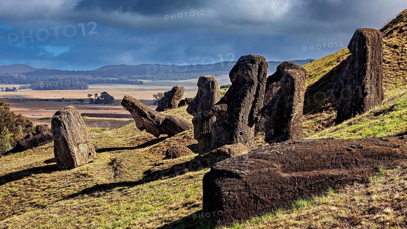 Moai statues lie and stand on a green hill under cloudy sky, The Moai statues on Easter Island in the Pacific Ocean