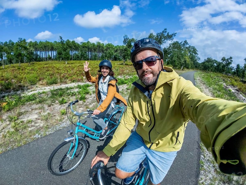 Happy couple cycling along a sunny pine lined path, taking a selfie together during an active holiday by a blue lake, smiling, waving and enjoying outdoor adventure and freedom
