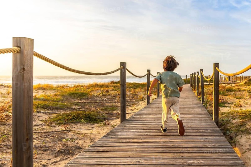 Little boy joyfully running along a wooden boardwalk towards the ocean as the sun sets, symbolizing freedom, adventure, and the idyllic essence of a childhood holiday at the beach