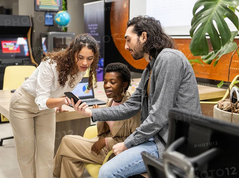 A diverse group of professionals collaborates in a modern office, discussing information displayed on a smartphone. The setting reflects teamwork, innovation, and workplace engagement