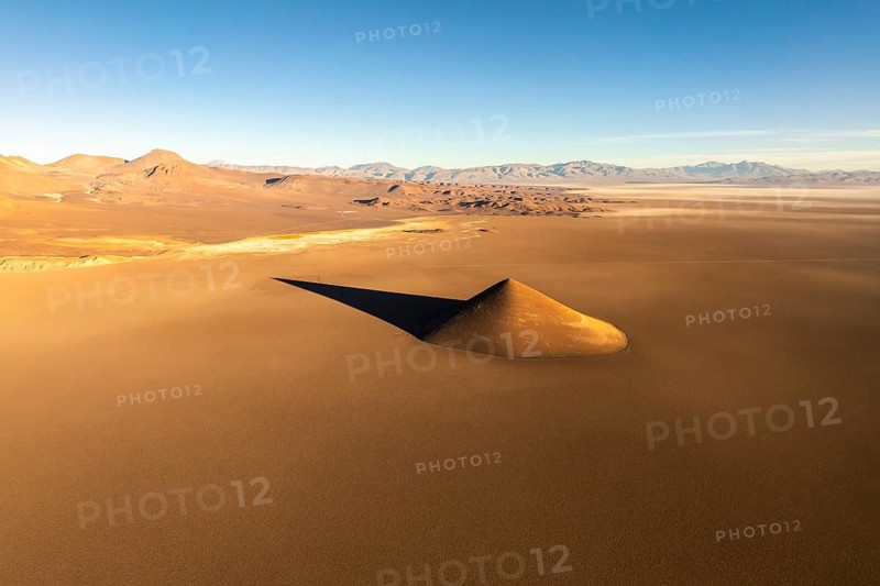 From above, the Cono de Arita rises dramatically from the flat puna landscape in Salta, Argentina. This natural volcanic formation presents a stark contrast against the vast, sandy expanse under the warm glow of the setting sun