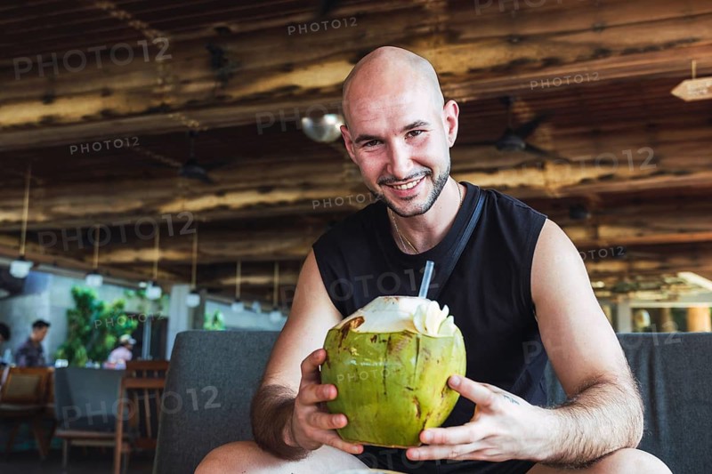 A cheerful bald man enjoys drinking a fresh coconut while relaxing in a rustic tropical cafe setting, embodying the spirit of vacation and holiday leisure in Bali