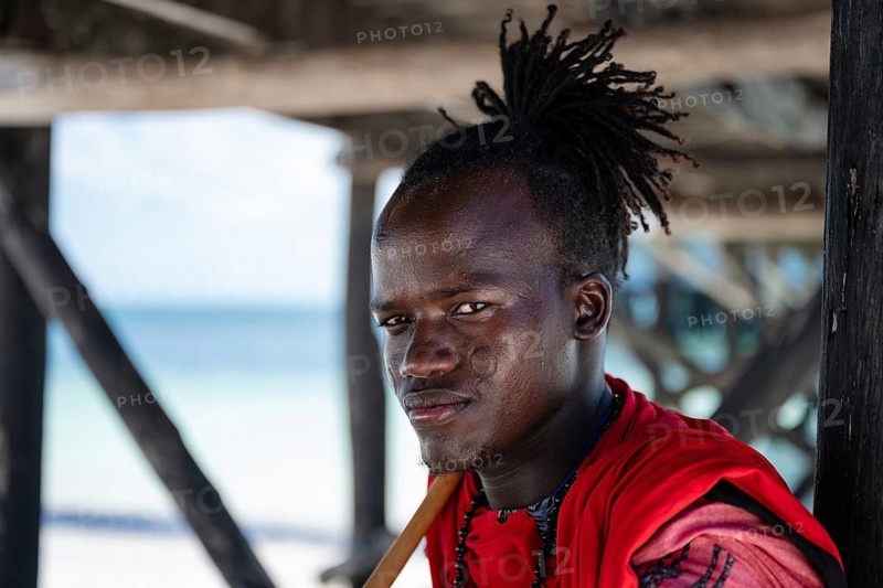 MICHAMVI, ZANZIBAR - OCTOBER 15, 2025: Close up portrait of a young African man in traditional red clothing looking at the camera with a beach background in Zanzibar, Tanzania