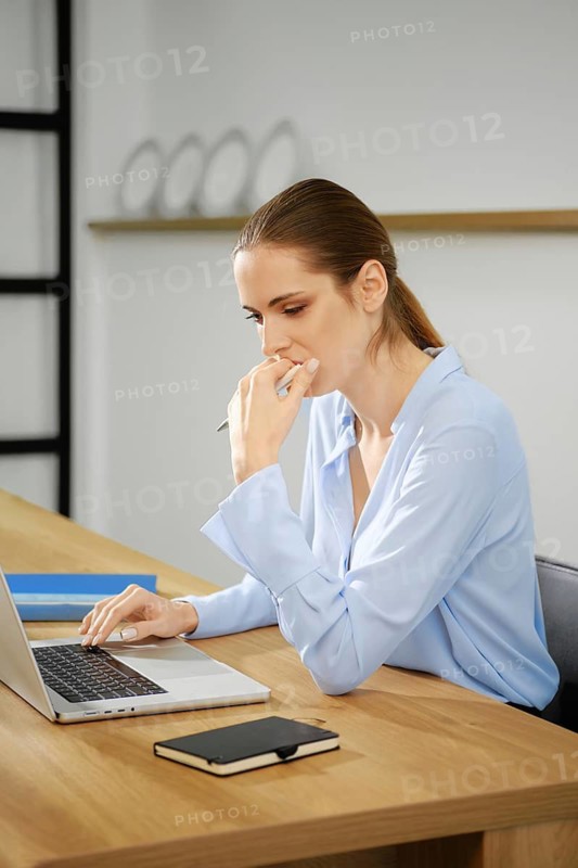 A young woman dressed in a light blue shirt is focused on her laptop while sitting at a wooden desk. She appears deep in thought, resting her chin on her hand in a contemporary office setting