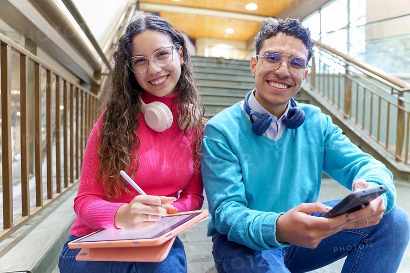 A cheerful young Gen Z woman with headphones and a tablet, and a man with headphones holding a smartphone, sit on stairs, embodying modern digital lifestyle and connectivity looking and smiling to the camera