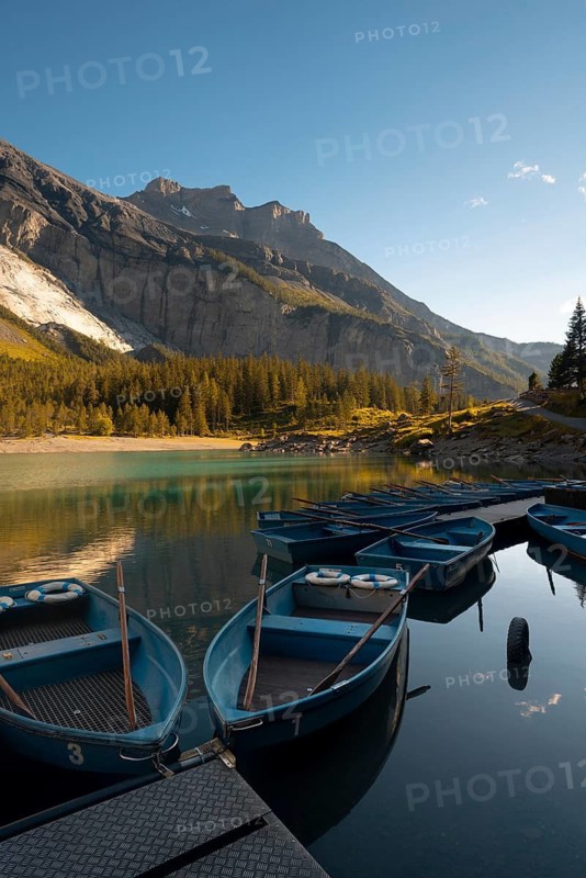 Scenic view of blue boats peacefully docked on the calm waters of Oachinensee, Switzerland, surrounded by lush greenery and dramatic mountain peaks under a clear sky