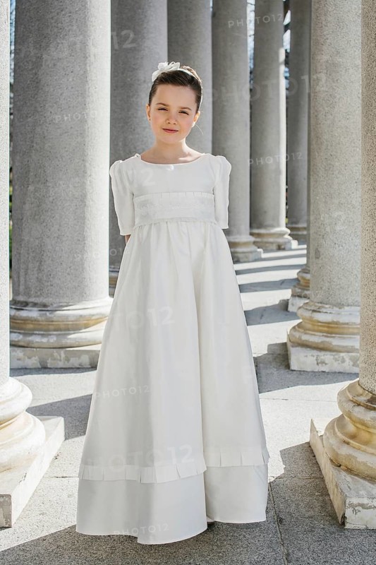 A young girl stands gracefully in a white First Communion dress, bathed in sunlight. She is surrounded by classical columns, exuding elegance and serenity