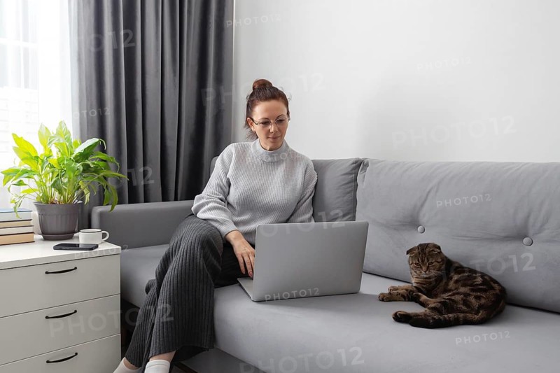 A woman in casual attire works on her laptop while sitting on a gray sofa next to a relaxed tabby cat. A plant and a cup of coffee are nearby, adding coziness