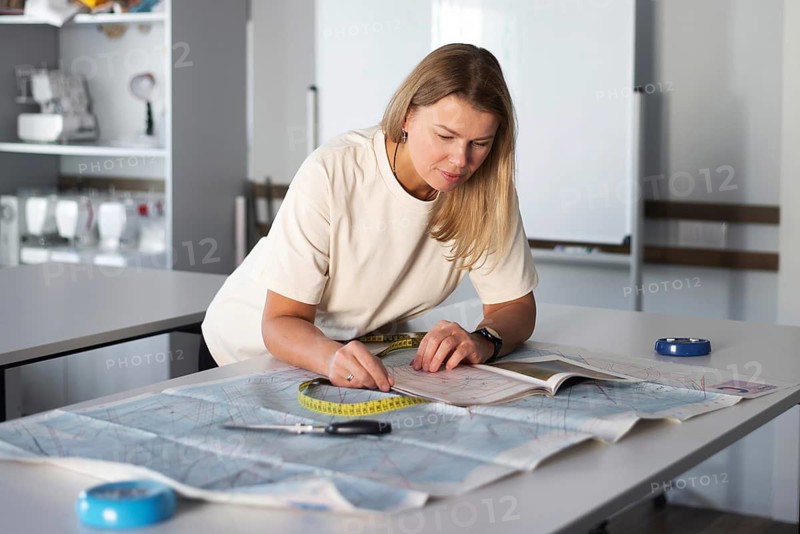 A woman is focused on her task in a sewing workshop, measuring fabric with a tape measure and working diligently. The room is filled with sewing equipment and patterns