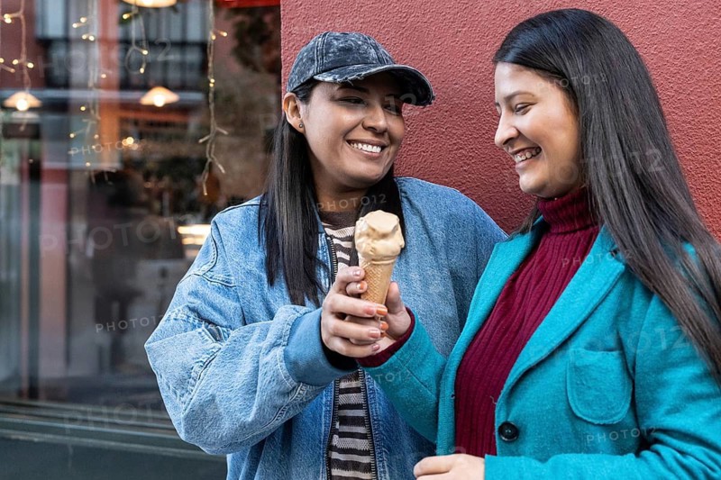 A joyful Latin lesbian couple shares an ice cream cone in a vibrant urban setting, showcasing affection and happiness. The scene is lively, with warm colors and smiles