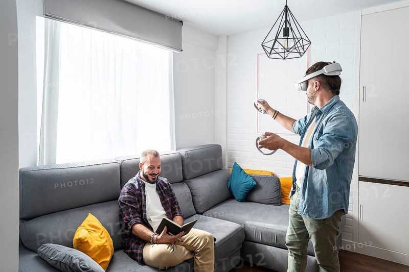 One man uses a VR headset, fully immersed in a virtual world, while his partner relaxes on the couch with a book. The cozy room invites relaxation and play