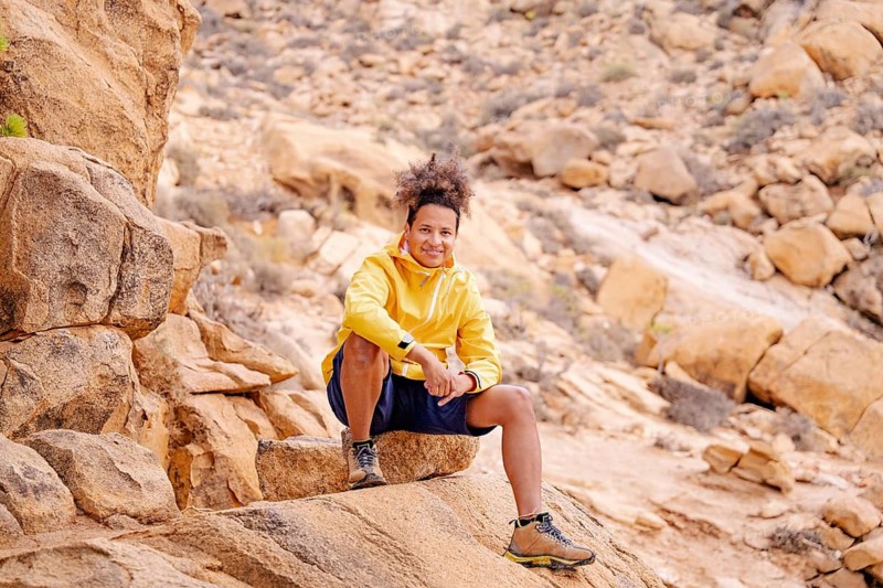 A young hiker in a yellow jacket sits on rugged rock formations of the Canary Islands, smiling confidently. The desert landscape showcases earthy tones, emphasizing adventure and outdoor exploration