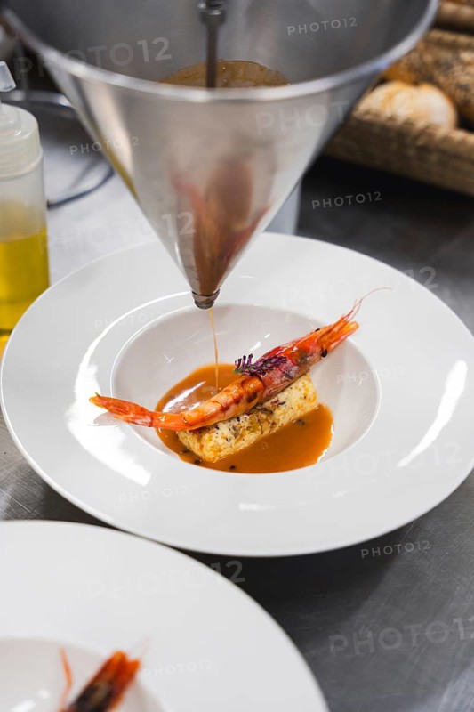 A chef precisely pours seafood sauce over a gourmet shrimp and terrine dish using a funnel, showcasing fine dining presentation in a professional kitchen setting