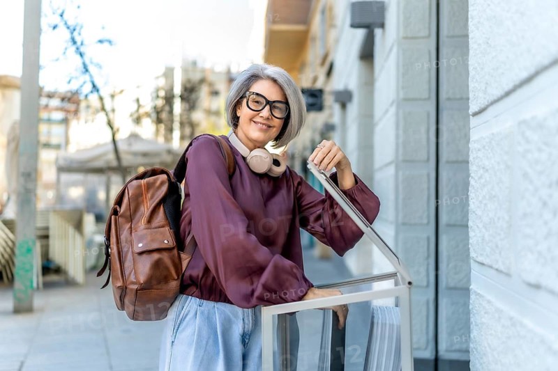 Woman with gray hair, glasses, and headphones wearing a backpack, smiling while pulling a piece of luggage along a city sidewalk, representing travel, urban exploration, and leisure