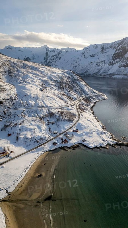 A breathtaking aerial view of a serene Nordic coastline, showcasing snow-covered landscapes, a meandering road, and a tranquil bay against a backdrop of majestic mountains, Kvaløya, Troms, Norway