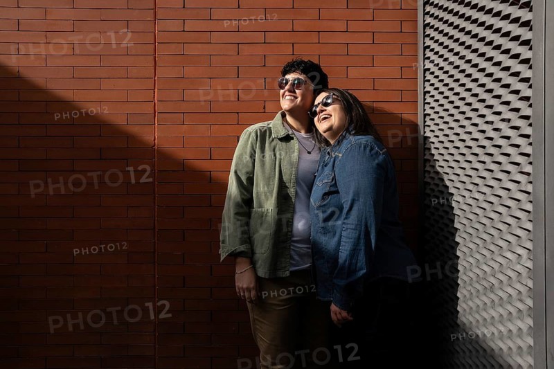 A curvy lesbian couple wearing sunglasses smiles together, leaning against a textured brick wall. They are enjoying each other company on a sunny day