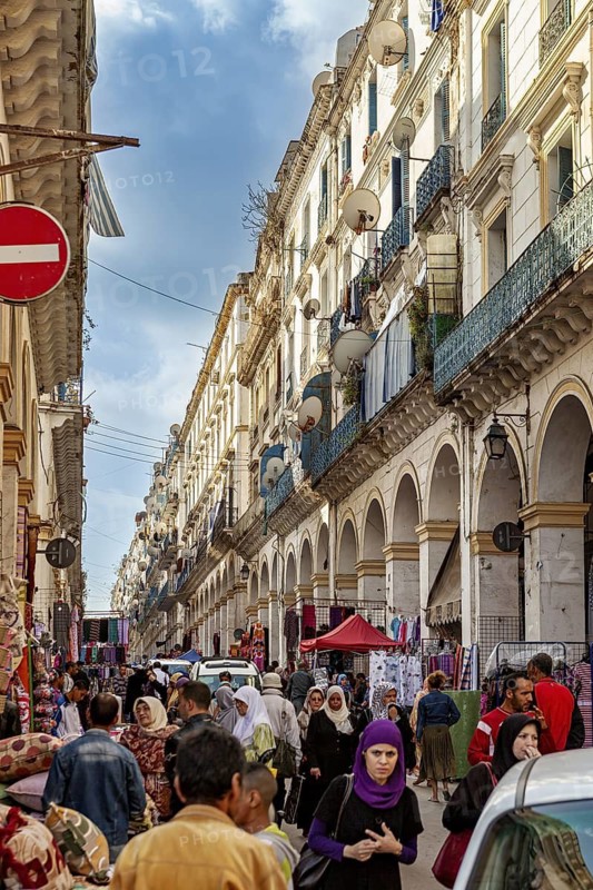 Busy street with many people and shops, surrounded by historic buildings with balconies under a blue sky, crowds of people in the old town of Algiers Algeria