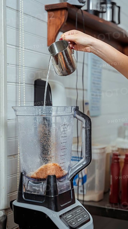 Hand pouring water from a metal pitcher into a blender filled with coffee grounds in a cozy cafe. Background includes shelves, bottles, and various kitchen utensils