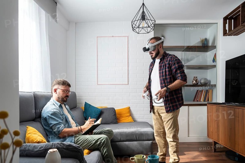 A gay couple engages in leisure activities in their living room. One reads a book while the other immerses in virtual reality gaming, creating a cozy and relaxed atmosphere