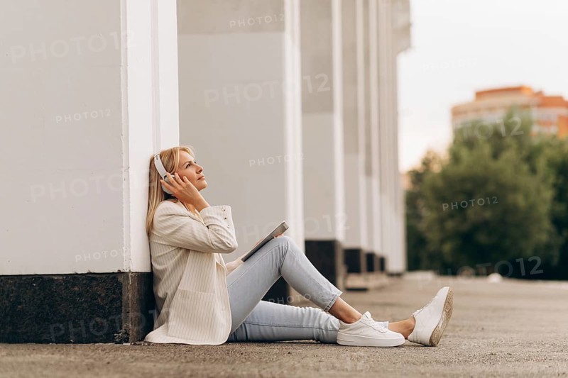 A businesswoman sits relaxed on the ground, listening to music through headphones. She leans back against a building wall, enjoying a peaceful moment outside in urban surroundings