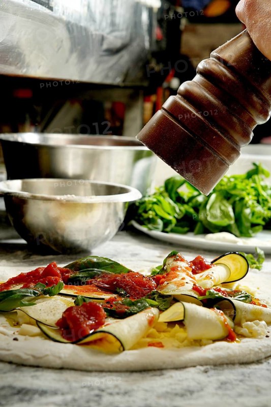 Cropped unrecognizable person seasons an uncooked pizza laden with fresh vegetables and cheese, enhancing the flavors before baking