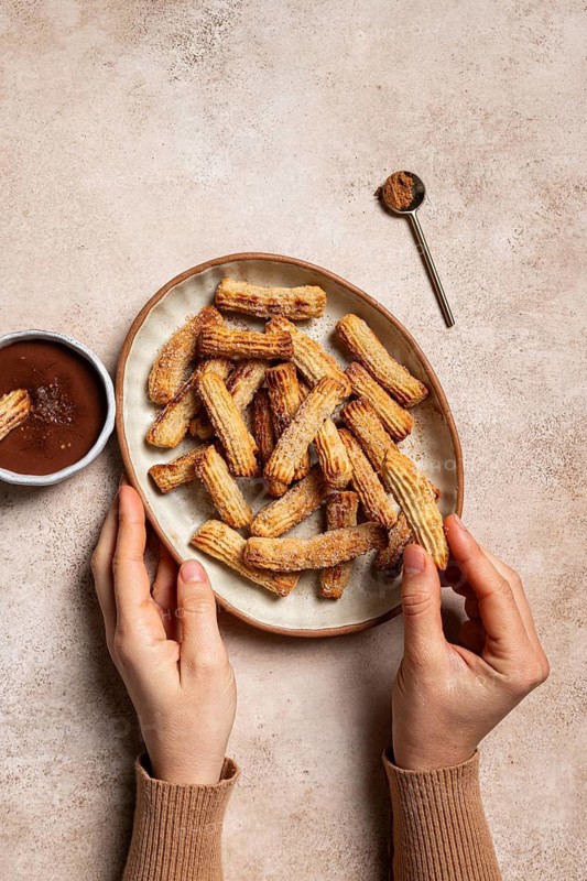 Top view of cropped unrecognizable hand on a ceramic plate filled with sugar and cinnamon coated churros, accompanied by a bowl of chocolate sauce