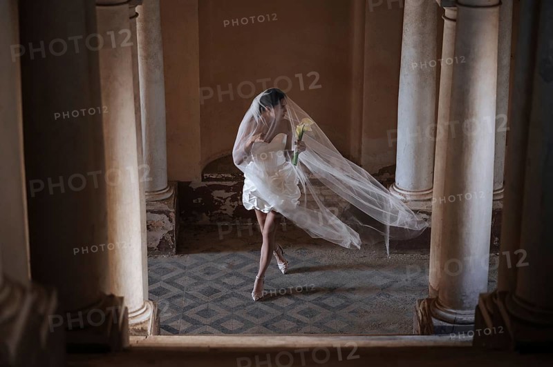 A bride in a flowing white dress and veil holds a bouquet while walking gracefully between columns in an ancient Italian villa, capturing the romance of a wedding in Treia, Macerata
