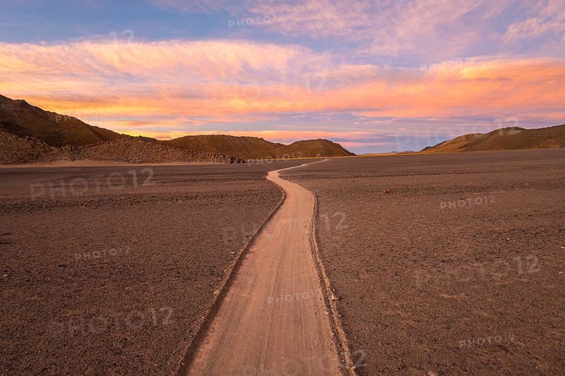 A tranquil desert scene features a winding path under a stunning sunset in the argentine Puna. The sky is painted with hues of pink and orange, creating a serene and surreal atmosphere