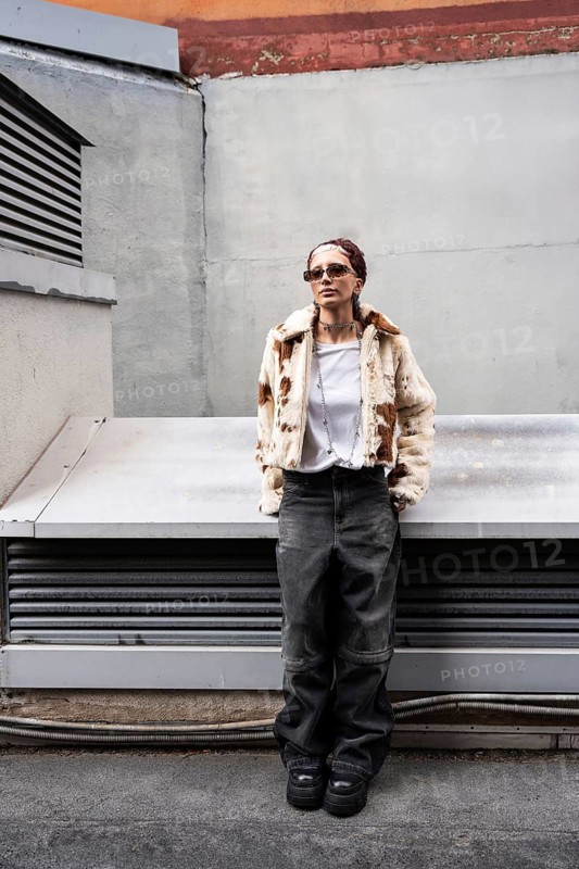 Stylish woman in sunglasses and fur jacket poses confidently against an urban backdrop. Casual white shirt and wide-leg jeans complete the contemporary streetwear look