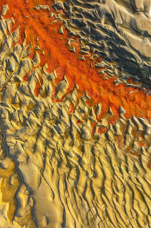 An aerial view reveals intricate sand patterns with vibrant orange and beige tones. The natural textures create a captivating abstract composition, resembling a painting