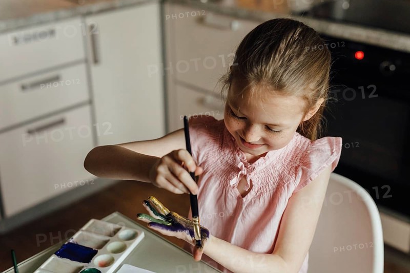 A young girl enthusiastically paints her hand using a brush while seated in a modern kitchen. She wears a pink blouse and is surrounded by art supplies, showcasing creativity