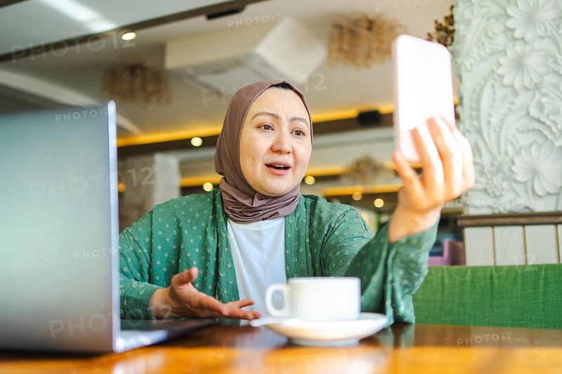 A Muslim woman in a hijab sits at a cafe table, engaging in a video call on her smartphone. A laptop is open beside her, with a cup of coffee on the table