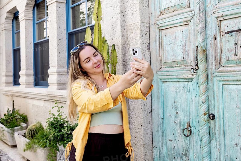 A female tourist joyfully takes a selfie on a vibrant street. Her yellow jacket contrasts with the rustic blue door, capturing a moment of travel excitement and exploration