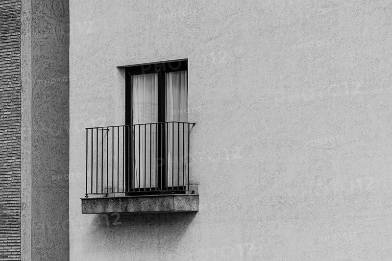 Black and white image of a minimalist exterior wall with a small balcony and a window. The textured surface of the wall contrasts with the sleek railing design, creating a modern aesthetic
