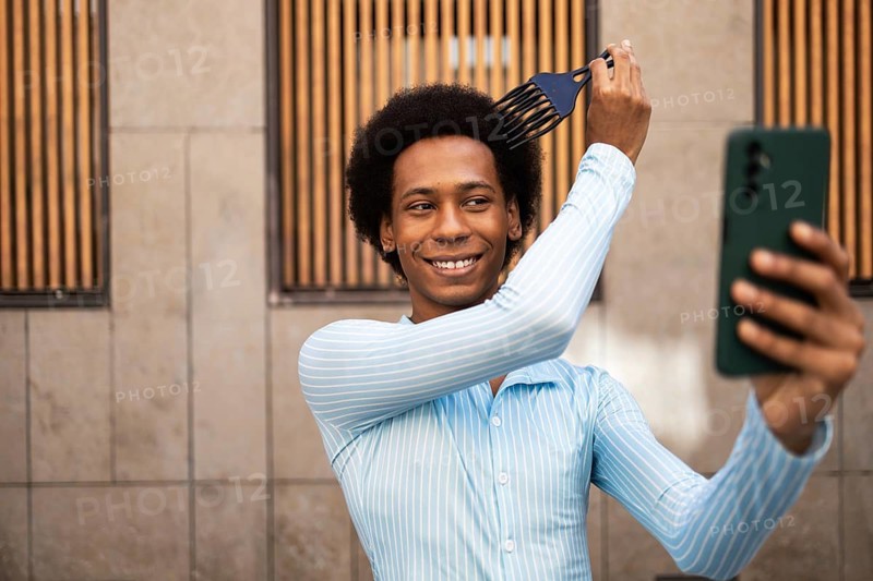 Smiling young non-binary adult styling hair with a pick comb while capturing a selfie. Wearing a blue shirt outdoors, they express joy and confidence in their appearance and identity