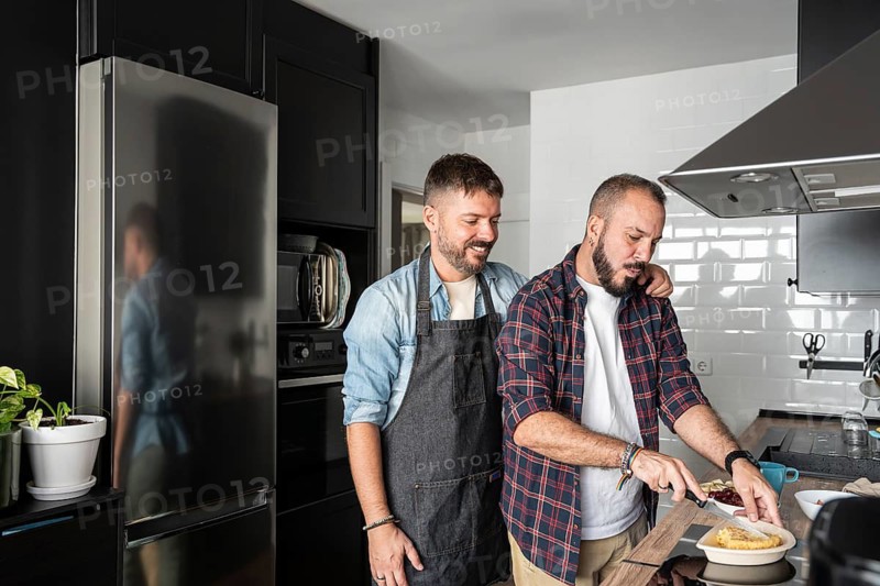 A loving gay couple prepares a meal together in their cozy, modern kitchen. One man embraces the other while he slices a dish on the counter, both smiling warmly