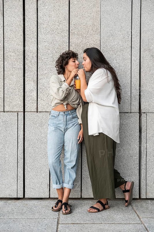 A lesbian couple shares a drink while leaning against a stylish concrete wall. They are enjoying a relaxed, tender moment together. Their casual attire adds to the scene's warmth