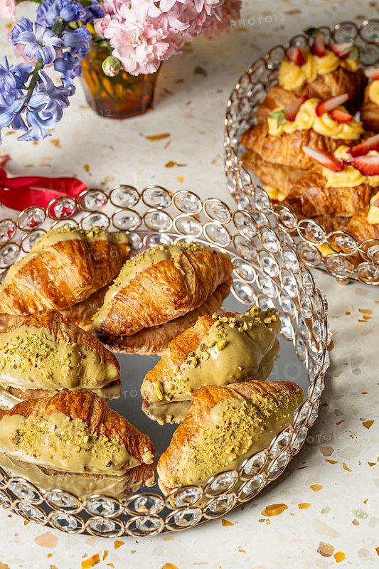 Elegant arrangement of croissants topped with pistachio cream, served on a crystal tray, accompanied by a vibrant floral bouquet on a stylish terrazzo table