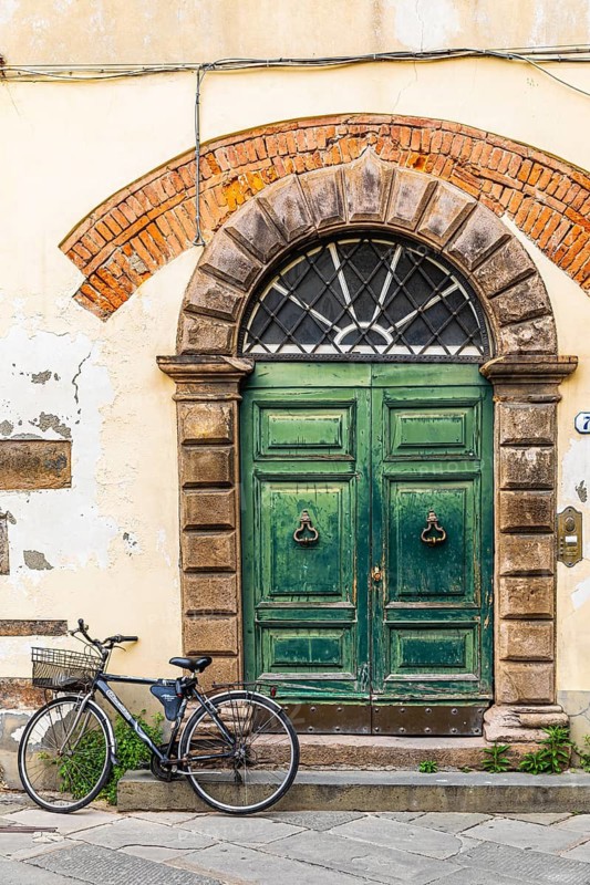 Weathered old arched entrance door, in front of it a parked bicycle, historic city centre, Lucca, Tuscany, Italy