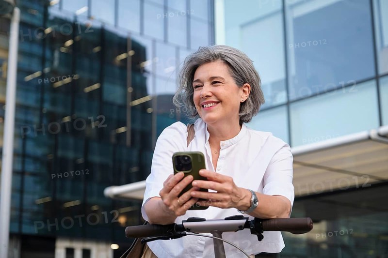 Mature woman smiling while holding a smartphone outdoors, dressed in a casual white shirt, enjoying leisure time in an urban setting with modern glass buildings in the background
