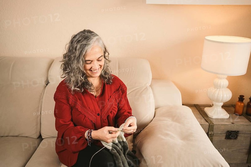 A woman with curly gray hair, wearing a red blouse, is knitting on a comfortable beige couch. A warm lamp light creates a relaxing atmosphere