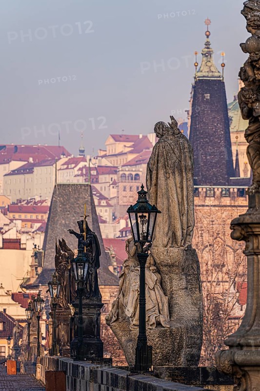 An evocative view of statues on Charles Bridge overlooking the historic rooftops of Prague during a calm sunset, capturing the serene ambiance and architectural beauty of the city