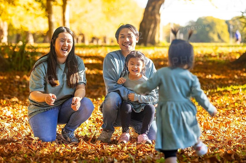 Joyful Asian family enjoying a playful afternoon in a scenic autumn park, surrounded by fallen leaves. The warm sunlight highlights their smiles as they embrace quality time together