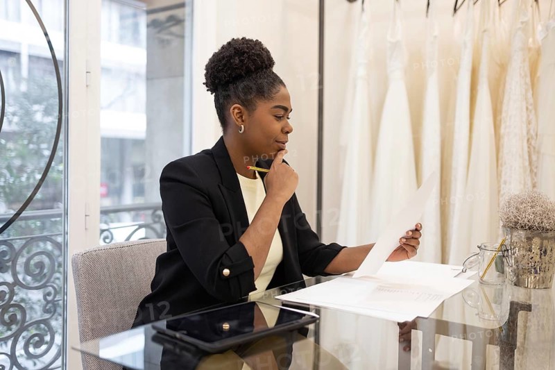Professional businesswoman in a bridal store reviewing documents attentively. Dressed in formal attire, she sits at a glass desk, surrounded by elegant decor and bridal gowns