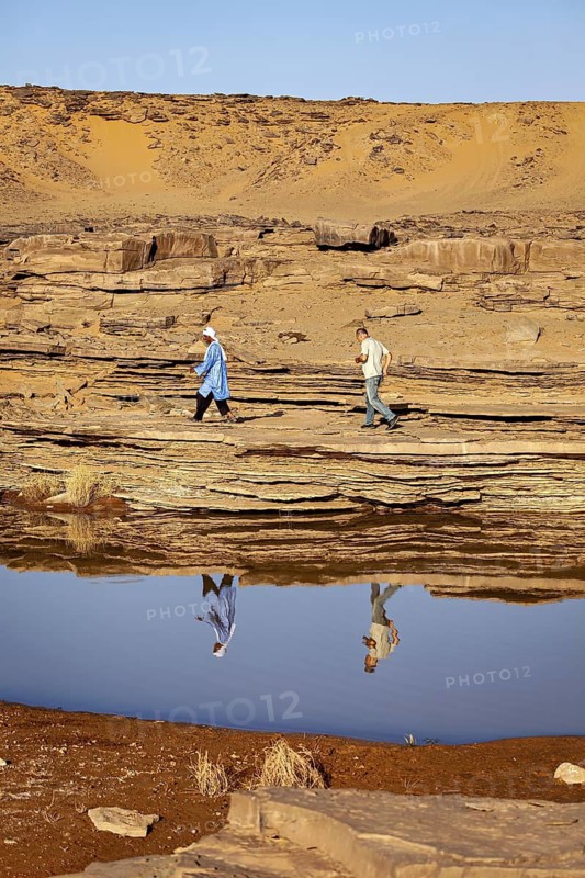 Two people walking over stone slabs in a vast desert landscape, Tuareg fetching water in the Sahara in Algeria