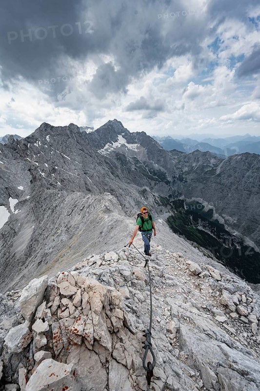 Mountaineer with helmet climbing on the rock, on a narrow rocky ridge, view of Zugspitze and Höllental in steep rocky mountain landscape, crossing the Jubiläumsgrat, ascent to Alpspitze via the southwest climb, Wetterstein range, Bavaria, Germany