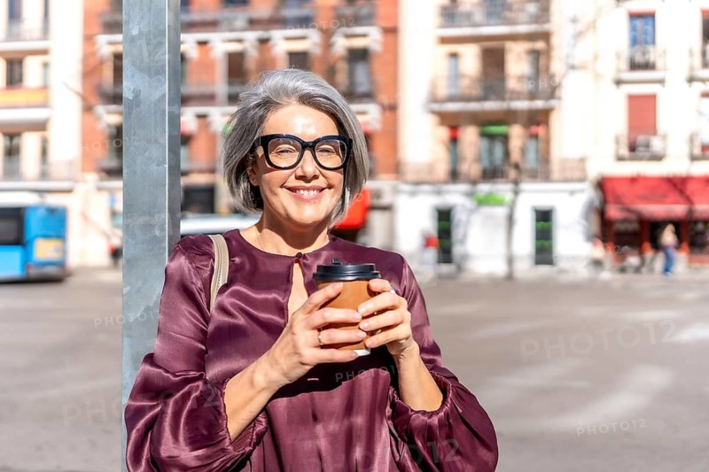 Mature woman with grey hair and eyeglasses smiling and holding a takeaway coffee cup while standing in a sunny european city square, confident and stylish commuter enjoying a break