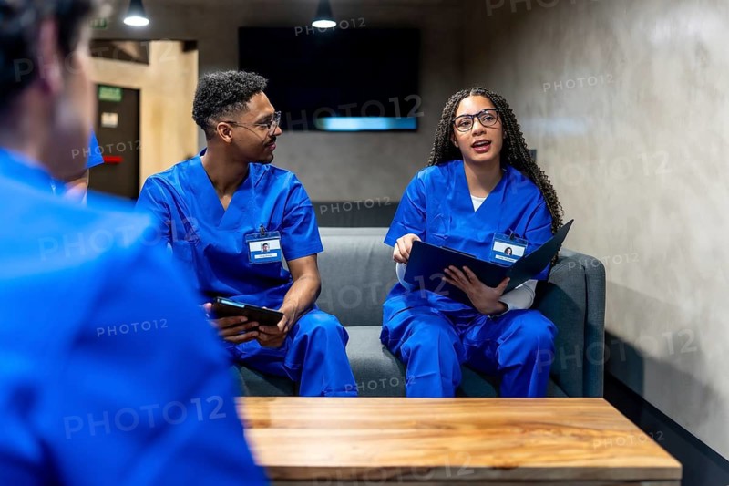 Healthcare professionals wearing blue scrubs and identification badges are discussing notes and information, engaging in a collaborative conversation while sitting on a sofa in a modern waiting area