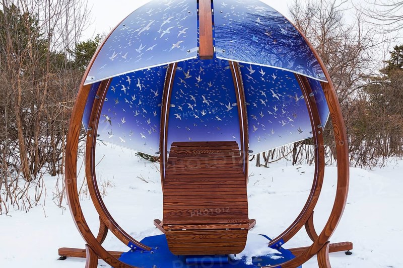 Interactive cocoon rocking chair made of curved wood and backlit blue tinted plexiglass panels canopy decorated with flock of birds in flight theme motifs created by Olivier Landreville and installed in winter like snow in early spring, Horizon Art Installation, Ile des Moulins, Old Terrebonne, Quebec, Canada