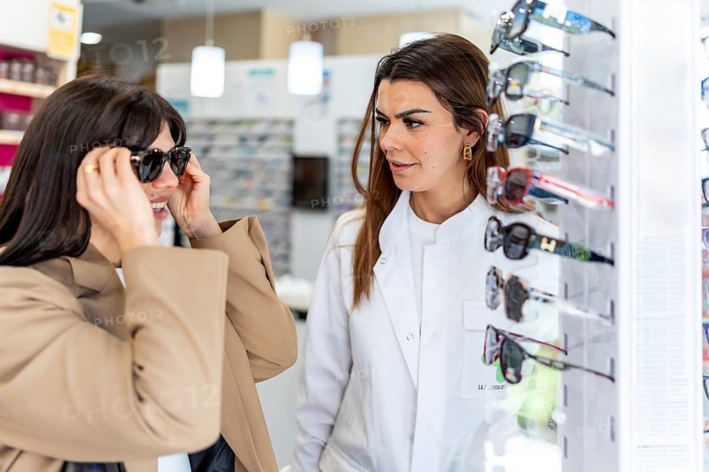 Woman trying on stylish sunglasses for better vision and eye protection, receiving professional assistance from an optician in a modern retail setting